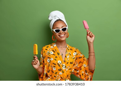Carefree positive dark skinned woman holds delicious ice cream, popsicles on stick, has fun during summer time, wears stylish sunglasses, yellow robe, wrapped towel on head, has sweet tooth.