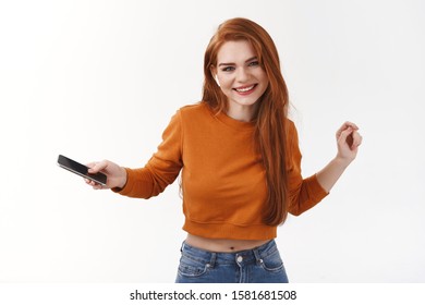 Carefree Pretty Caucasian Redhead Woman In Orange Cropped Sweater, Holding Smartphone, Dancing And Moving Rhythm Music, Listen Catchy Song In Wireless Earphones, Smiling Camera Happy