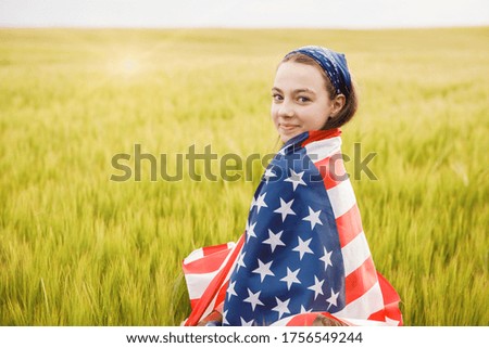 Carefree pretty girl running and smiling on the green field with a blowing flag of USA. Independence Day