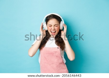 Carefree pretty girl scream from joy, listening music in headphones with happy face, lip-sync lyrics, standing over blue background