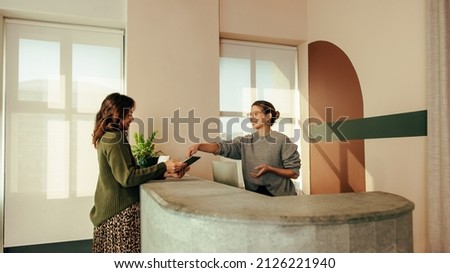 Carefree receptionist assisting a woman with signing in to an office. Friendly receptionist showing a woman where to sign on a digital tablet. Woman working at the front desk of a co-working space.