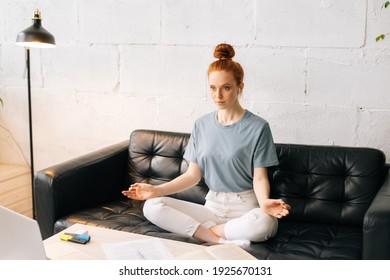 Carefree Redhead Young Woman Takes Deep Breath, Practices Yoga Or Meditation, Sitting On Sot Couch With Crossed Legs. Lady Sitting In Lotus Pose At The Desk With Open Eyes.