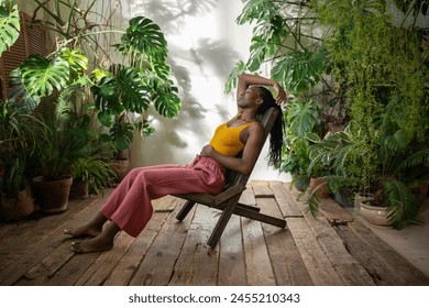 Carefree relaxed african american woman take break surround by tropical indoor plants. Satisfied young black female rest sits barefoot with closed eyes on chair in urban jungle home with houseplants. - Powered by Shutterstock