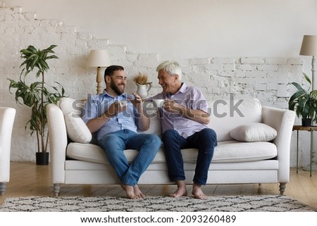 Carefree relaxed sincere happy two male generations family, young son and elderly senior father involved in pleasant conversation, talking sharing life news, drinking hot tea or coffee at home.