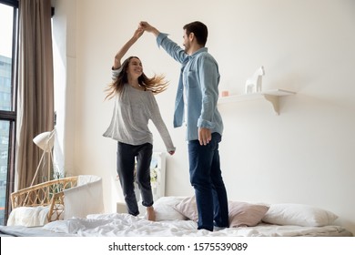 Carefree romantic young couple having fun dancing on bed at modern home, happy active husband and wife enjoying funny party on honeymoon together on leisure morning lifestyle in bedroom interior