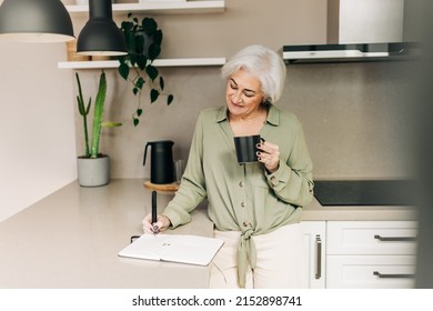 Carefree Senior Woman Writing Notes In Her Diary While Standing In Her Kitchen At Home. Happy Mature Woman Making A To-do List While Enjoying A Cup Of Coffee.