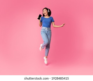 Carefree Singer. Asian lady singing favorite song and using cell phone as a mic, dancing and jumping, wearing wireless headphones. Excited woman having fun at pink studio, enjoying sound, full length