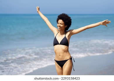 Carefree smiling African American female traveler in swimwear strolling with raised arms on coast along sea in tropical resort on summer day and enjoying freedom