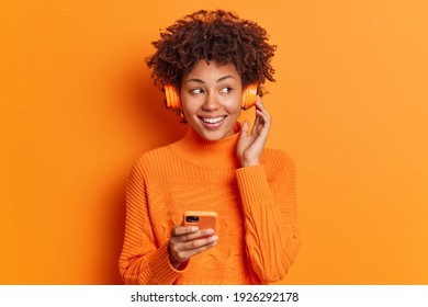 Carefree smiling dark skinned young curly woman listens music in wireless headphones holds mobile phone chooses song from playlist dressed in casual sweater isolated over vivid orange background. - Shutterstock ID 1926292178