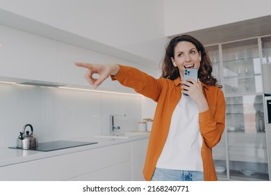 Carefree Spanish Woman In Earphones Is Singing With Cellphone As With Microphone. Application For Music Listening Online. Playful Passionate Girl Is Singing At Home And Points To You.