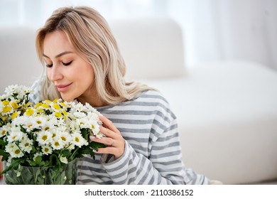 Carefree stylish good-looking woman ordered flowers for home, close eyes smiling and sniffing beautiful bouquet of wildflowers, sitting satisfied in bright cozy living room. portrait