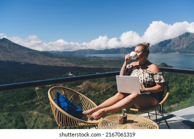 Carefree Swedish woman sitting on hotel balcony with laptop computer while working remote on freelance. tattooed female digital nomad enjoying work on netbook and drink coffee, mountains view