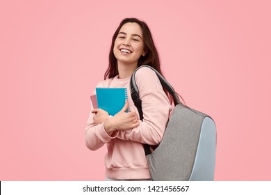 Carefree teen girl with backpack smiling and embracing notepads during studies against pink background
