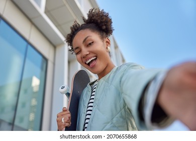 Carefree teenage girl winks eye and poses for making selfie hold penny board practices different skateboard tricks feels cool poses outdoors near building against blue sky. Youth and active rest - Powered by Shutterstock