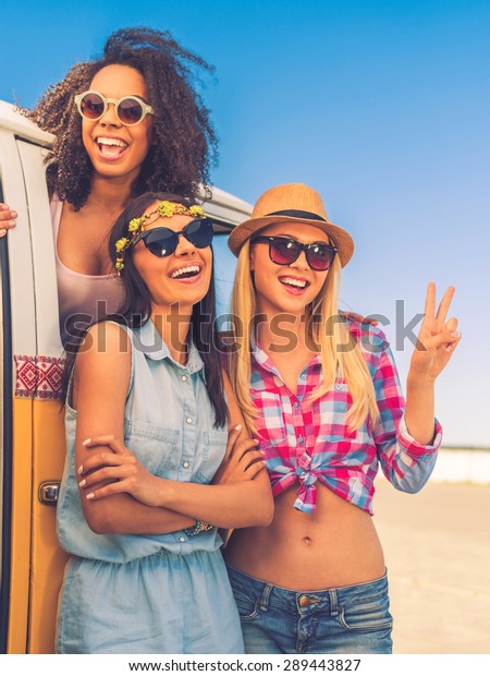 Carefree time with friends. Happy young African woman\
looking out from the retro minivan while two her friends standing\
near her