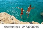 Carefree tourists hold hands while jumping into the refreshing blue sea during a relaxing summer vacation. Active young woman and her boyfriend dive off a high cliff and into the deep blue ocean.