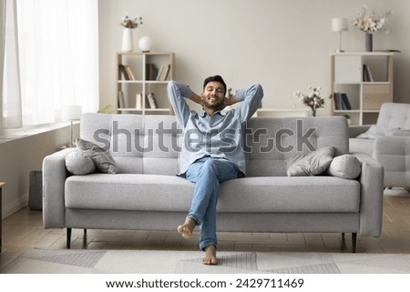 Carefree wellbeing barefoot Indian guy enjoy fresh conditioned air leaned on comfortable sofa in fashionable living room, relaxing alone with hands behind head. Rest, leisure, daydreams, smart home