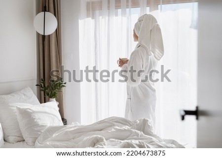 Carefree woman with coffee cup in hands standing near window in hotel room. Female in white bathrobe enjoying weekend day in apartment, resting after beauty procedure. Advertising of resort and spa