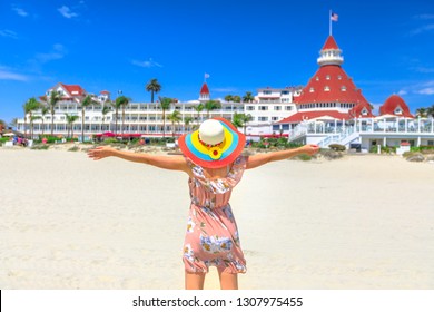 Carefree woman in hat on Coronado Central Beach, looking historic Victorian hotel on Coronado Island, San Diego. Lifestyle tourist enjoying ummer season in California West Coast, USA. Sunny blue sky.