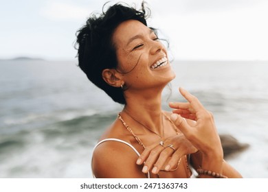 Carefree woman with wavy hair and bohemian jewelry posing joyfully by the seaside beach, expressing joy and peace. - Powered by Shutterstock