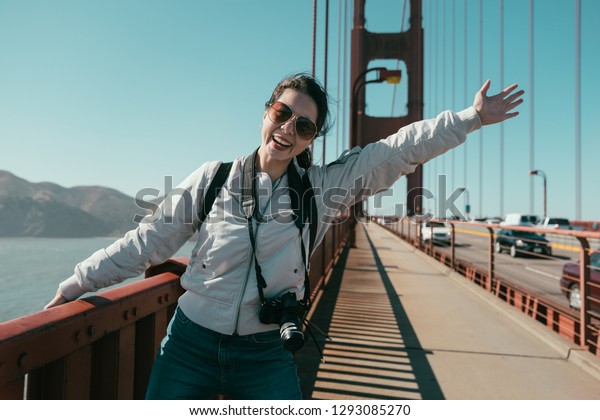 carefree young asian girl standing on golden state\
bridge face camera cheerful laughing raising hands arms showing\
sharing beautiful view with blue sky in summer. woman enjoy nature\
sightseeing sea.