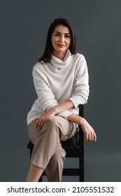 Carefree young businesswoman sitting on a chair, dressed in white sweater and beige pants, posing in studio over gray background. Relaxed and beautiful young woman