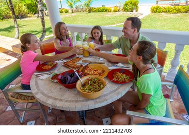 Carefree Young Caucasian Family Having Fun Toasting Juice And Enjoying Eating Dinner At Exotic Outdoor Beach Restaurant Caribbean