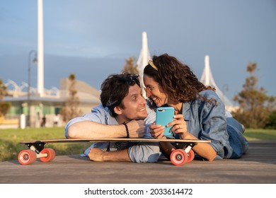 Carefree young couple surfing internet or shopping online outdoors using mobile phone internet lying in city park on longboard look at each other with playful cheerful smile. Urban lifestyle concept - Powered by Shutterstock