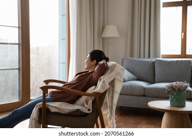 Carefree Young Dreamy Indian Woman Relax On Comfort Armchair With Hands Behind Head Looking Out Window Enjoy Day Off In Cozy Modern Living Room. Serenity, No Stress, Weekend At Home, Daydreams Concept