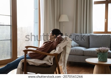 Carefree young dreamy Indian woman relax on comfort armchair with hands behind head looking out window enjoy day off in cozy modern living room. Serenity, no stress, weekend at home, daydreams concept