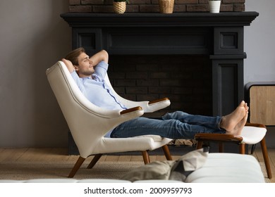 Carefree Young Male Homeowner Relaxing Barefoot In Capacious Armchair At Luxury Apartment By Modern Fireplace. Serene Single Millennial Male Rest With Hands Behind Head At Rich Comfortable Living Room