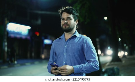 Carefree Young Man Walking In Drizzle Rain Outside At Night In City Street Sidewalk Holding Phone. Happy Guy Walks Outdoors