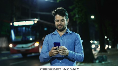 Carefree Young Man Walking In Drizzle Rain Outside At Night In City Street Sidewalk Holding Phone. Happy Guy Walks Outdoors