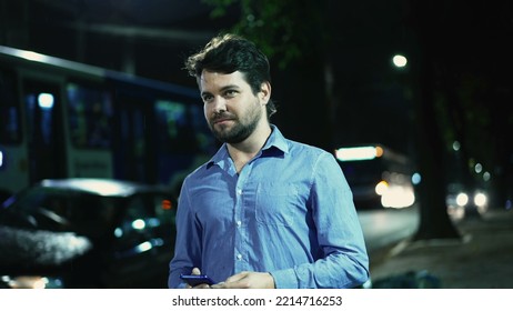 Carefree Young Man Walking In Drizzle Rain Outside At Night In City Street Sidewalk Holding Phone. Happy Guy Walks Outdoors