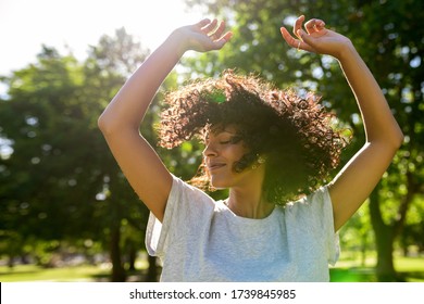 Carefree young woman dancing with her arms raised outside in a park on a sunny summer afternoon twirling her curly hair