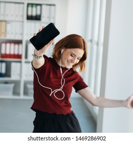 Carefree young woman dancing to music in the office as she listen on ear buds to tracks on her mobile phone - Powered by Shutterstock