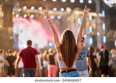 Carefree young woman enjoys summer music festival on beach, raises peace sign hands, celebrating with crowd. Sunset lights up stage, audience engrossed in concert. Casual, fun, youth lifestyle.