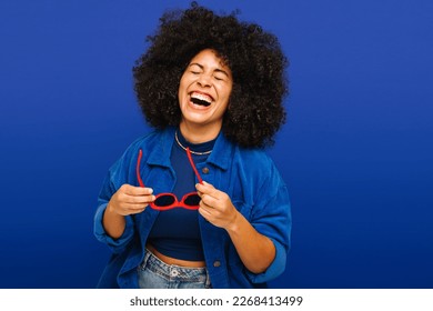 Carefree young woman laughing happily while holding sunglasses in a studio. Fashionable woman with curly hair having a good time while standing against a blue background. - Powered by Shutterstock