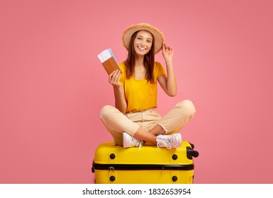 Carefree Young Woman In Summer Hat Holding Passport And Tickets, Sitting On Big Yellow Suitcase, Ready To Travel. Cheerful Lady Traveller Posing Over Pink Studio Background, Ready For Vacation