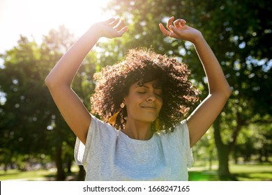 Carefree Young Woman Twirling Her Curly Hair While Dancing With Her Arms Raised Outside In A Park On A Sunny Summer Afternoon