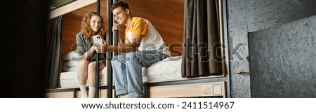 carefree youthful woman showing smartphone to boyfriend on double-decker beds in hostel room, banner