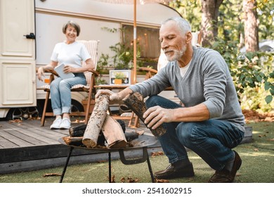 Careful senior old elderly husband laying a fire with firewood while wife relaxing on the porch of camper van motor home trailer. Adventures ahead concept - Powered by Shutterstock