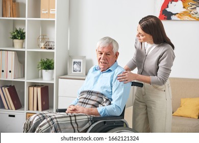 Careful young nurse talking to elderly man in wheelchair while visiting him at home