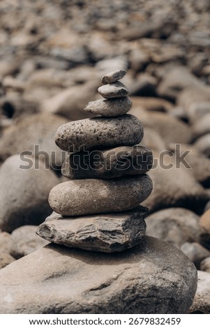 A carefully balanced stack of smooth river stones in varying sizes and textures, arranged in perfect equilibrium against a blurred rocky shoreline background. The weathered stones showcase natural ear
