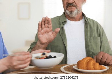 Caregiver with breakfast and elderly man refusing to eat it at wooden table indoors, closeup