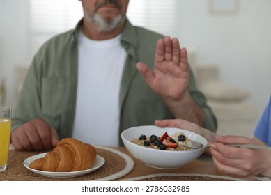 Caregiver with breakfast and elderly man refusing to eat it at wooden table indoors, closeup