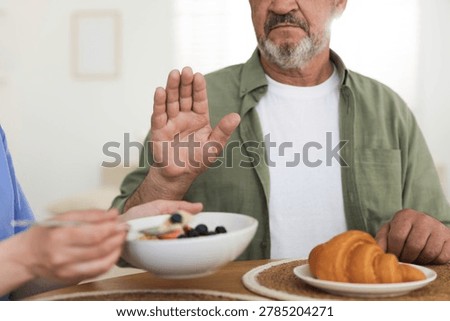 Caregiver with breakfast and elderly man refusing to eat it at wooden table indoors, closeup