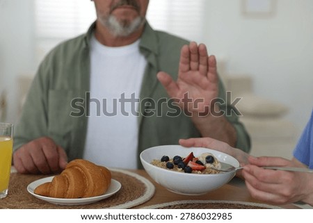 Caregiver with breakfast and elderly man refusing to eat it at wooden table indoors, closeup