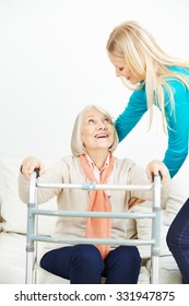 Caregiver Helping Old Woman With Walker Getting Up From Sofa At Home