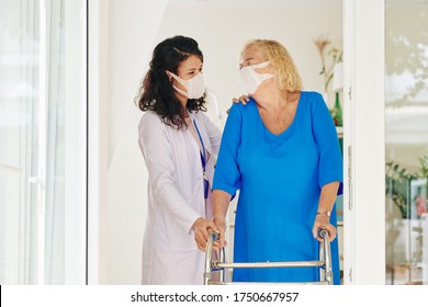 Caregiver In Medical Mask Helping Senior Woman To Move With Help Of Walker